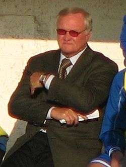 A man sitting in a dugout with his arms folded during a football match. He is wearing a pair of sunglasses as he is looking into the sun