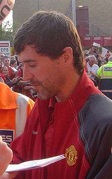 A man with dark hair and stubble, wearing a red jacket with grey sleeves.
