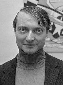 A black-and-white portrait photograph of a man at bust length facing and looking at the viewer while wearing a turtleneck shirt under a blazer all in front of a painting