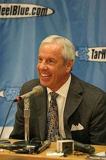 A man with short gray hair wearing a suit smiles in front of a microphone