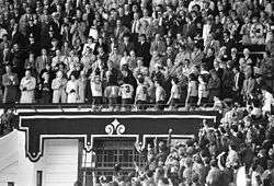 A man stands in the Royal Box lifting a trophy with his teammates walking behind him. A large crowd looks on.