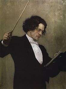 A middle-aged man with long dark hair, wearing a tuxedo and standing behind a music stand, waving a conductor's baton.