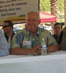 A smiling, somewhat heavy-set man in his sixties with thinning white hair and wearing a green and blue floral print shirt sits behind a table under a covering outdoors. On his shirt is a sticker saying no to tax hikes and in front of him is a bottle of water. Several other men are sitting near him; in the background is a sign with a quotation about liberty from John Adams, some cars in a parking lot, and tall palm trees.