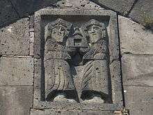 Two bearded figures in stone relief, holding an object that looks like an open door with a bell on top.