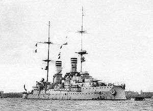A large light gray warship sits motionless in harbor; it has two tall smoke stacks, both painted with three dark bands for identification.