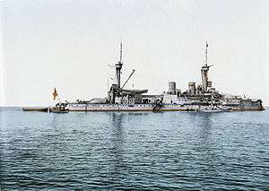 A large light gray battleship with two tall masts sits motionless in calm water. Several smaller boats are tied alongside.