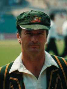 Person aged around 30 wearing a baggy green cap with the Australian coat of arms, Australian blazer, green with yellow stripes, and a cream cricket shirt. He is clean shaven and has brown hair.