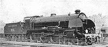 Side-and-front view of a 4-6-0 locomotive on an turntable. A member of the crew is standing next to the front.