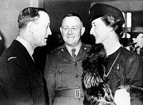 Two uniformed men with a woman in a dark dress, hat and fur stole