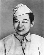 Head and shoulders of a smiling young man wearing a garrison cap and a shirt and tie, the bottom of the tie tucked into the shirt between the buttons.