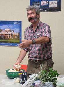 A color image of Sandor Katz, a man in a loosely worn purple plaid shirt and cargo pants with grey hair and two shade of grey mutton chop sideburns and mustache. He is standing at a table in which he is making sauerkraut. He has one hand in the bowl and is making a circular gesture with the other.