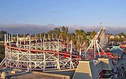Evening photograph of the white, wooden Santa Cruz Roller Coaster, amusement park structures, and a background of eucalyptus and palm trees.
