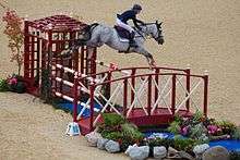 A gray horse, ridden by a woman, in mid-air over a red and white striped fence.
