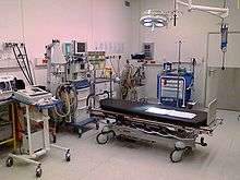 Color photograph of a room designed to handle major trauma. Visible are an anesthesia machine, a Doppler ultrasound device, a defibrillator, a suction device, a gurney, and several carts for storing surgical instruments and disposable supplies.