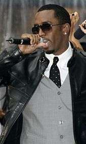 A man wearing a white dress shirt, tie, gray vest, black jacket, and sunglasses, singing into a microphone.