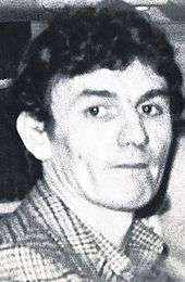 A black-and-white photo of a white male with black hair, wearing a chequered shirt
