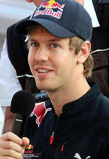 A man in his early twenties wearing a navy blue baseball cap with sponsors logos and a T-shirt jacket. He is holding a microphone in his right hand.