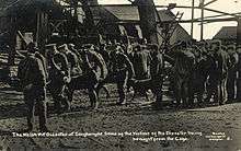  A uniformed stretcher party carry a body away from the pithead; others wait around the pithead. Wreckage from the explosion can be seen in the background.