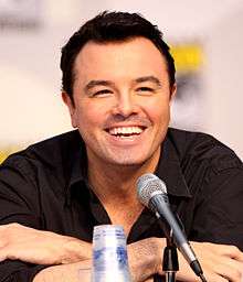 A man with black hair, and tan skin with a black shirt on, leans forward while laughing into a microphone.