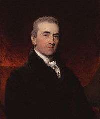 an older gentleman with short, greying hair, although his eyebrows remain black. He is wearing a black jacket with a small white cravat, and is sat in front of some dark red paper or cloth.