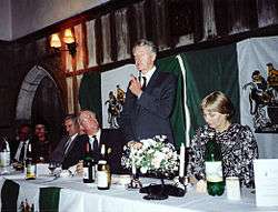 Photo of smartly dressed dinner guests at a long table watch standing man talk. Near camera sits a middle-aged woman with chesnut hair cut short.