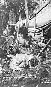 Black and white photo of a woman kneeling on the ground making a snowshoe. She is wearing a plaid shirt and white dress locking down at the snowshoe. Around her is four frames of snowshoe to be made leaning on a tippy,