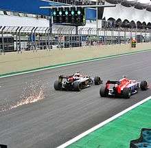 A picture a Lotus E23 Hybrid and a Marussia MR03B driving side-by-side during the 2015 Brazilian Grand Prix, with sparks flying up from behind the Lotus.