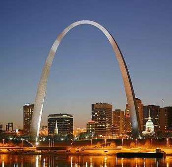 Night view of a city skyline, with skyscrapers in the background, a towering arch in the center and a river in the foreground