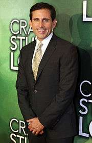 The image is of actor Steve Carell. He is standing in front of a green background, wearing a suit, and smiling.