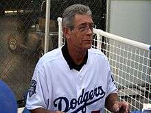 A gray-haired white male, wearing a white uniform with "Dodgers" across it, sitting in a bullpen with a white fence in the background.