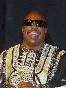 A man smiling, wearing black sunglasses and colorful clothing, behind a microphone.