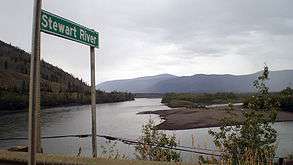 A wide river flows through hills. A sign in the foreground says "Stewart River".
