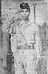 A man, saluting; he is wearing a military uniform and peci.