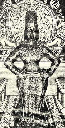 A black-and-white image of an idol of an arms-akimbo bare-chested man, wearing a conical head-gear, a dhoti and ornaments. The idol is placed on a brick, and backed by a decorated halo.
