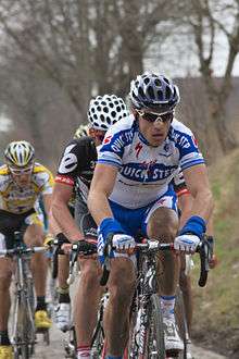 A road racing cyclist wearing a white and blue jersey with red trim and sunglasses leads a line of other cyclists. Partially visible behind him are a cyclist in a black and white jersey and one in a white and yellow jersey.