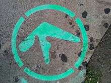 The edge of the pavement on a city street in New York, United States. Green-coloured spray-painted graffiti depicts the Aphex Twin logo inside a dashed outer circle. At the bottom right is roadside greenery.