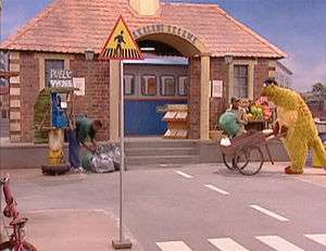 Television set, showing a large brick building on a city street; to the right, a large puppet is pushing a wheelbarrow and to the left, a man, next to a phone booth, is bending over several mail bags. Closer to the front of the image is a signpost, with a triangle that has a black figure and a yellow background, on top.
