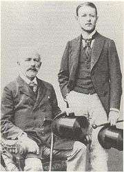 Two men in dark jackets, waistcoats and light slacks and carrying top hats and canes, a man in his 20s with dark hair standing and the second in late middle age with balding grey hair and a beard