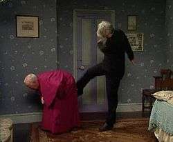 A priest, dressed in black, kicks a bishop in a purple cassock in the backside as he bends down.
