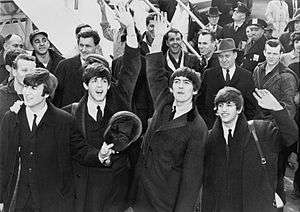 A black-and-white image of Paul McCartney, George Harrison, John Lennon and Ringo Starr waving to fans after arriving in America in 1964. A crowd is visible behind them on the left.