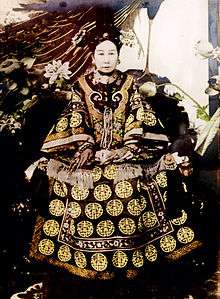 Full-face black-and-white photo of a woman with long fingernails sitting on a throne, wearing a richly adorned robe, a complicated hairdo, and a multi-layered pearl necklace. There are different kinds of flowers around her, as well as what appear to be peacock feathers.