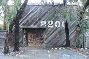 Large, wooden building with a brown door (showing woodland animals play musical instruments) located in the bottom, centre left, and the large numbers "2200" painted in white above the door, centre-right. Asymmetrical trees with hanging foliage frame the building on all sides, while on the asphalt in the foreground, there are parking spaces and a disabled person sign.