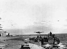 Black and white photograph of a single engined monoplane flying just above the deck of an aircraft carrier with its wheels extended. Two other aircraft are visible flying in the background of the photo.