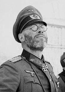 A black-and-white photograph of a man wearing a military uniform and a neck order in shape of an Iron Cross. He is wearing glasses and is looking up into the sky.