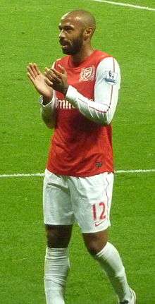 A shaven-headed man in a red football shirt with white sleeves, shorts and socks, applauding the crowd.