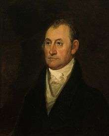 A man with receding, dark hair wearing a high-collared white shirt, a vest, and a black jacket