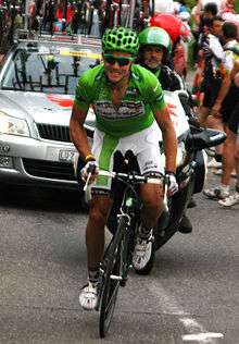 A cyclist in a green jersey bearing the Cervélo logo. He also wears a green helmet and sunglasses with green frames, and has a pained look on his face as he climbs a hill on his own. Behind him are a car and a motorbike, and spectators watch from the roadside.