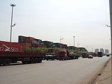 line of heavy trucks going north, background a container yard