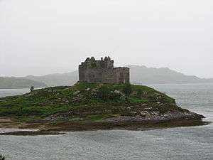 A small rocky islet with the ruins of a small medieval castle in the centre. Low rocky hills are visible in the background through the mist.