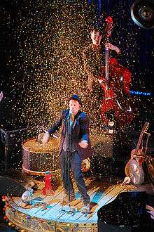 A man performing on-stage, surrounded by confetti. Several instruments surround him and behind him a man is playing a double bass.